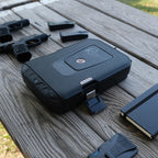 Portable biometric safe with keypad on outdoor table, next to handgun magazines and notebook