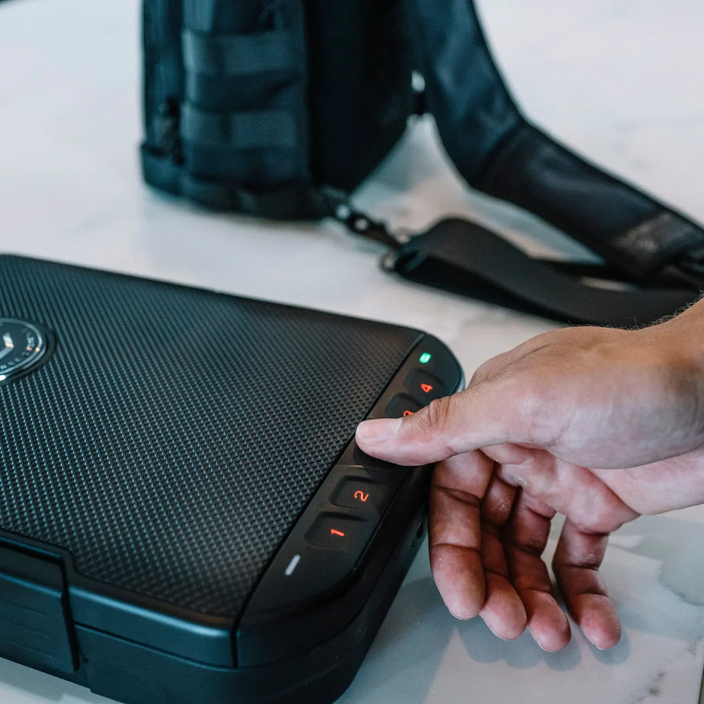 Person unlocking compact travel safe with keypad, black tactical bag in background