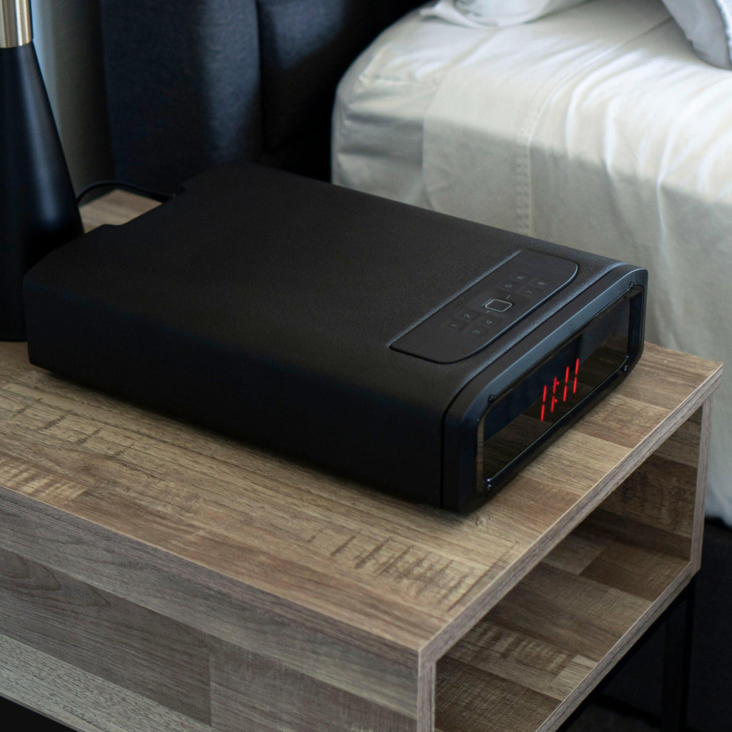 Black biometric safe with digital keypad and LED clock on a wooden nightstand beside a bed.