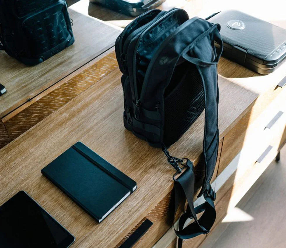Black tactical backpack on wooden desk next to tablet, notebook, and compact safe