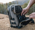 Tactical backpack on ground outdoors, open front pocket showing organized gear compartments