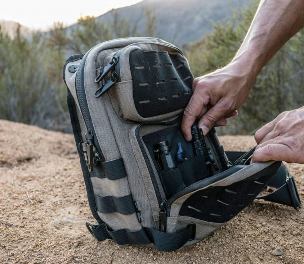 Hands opening a tactical backpack outdoors, revealing organized gear in rugged terrain.