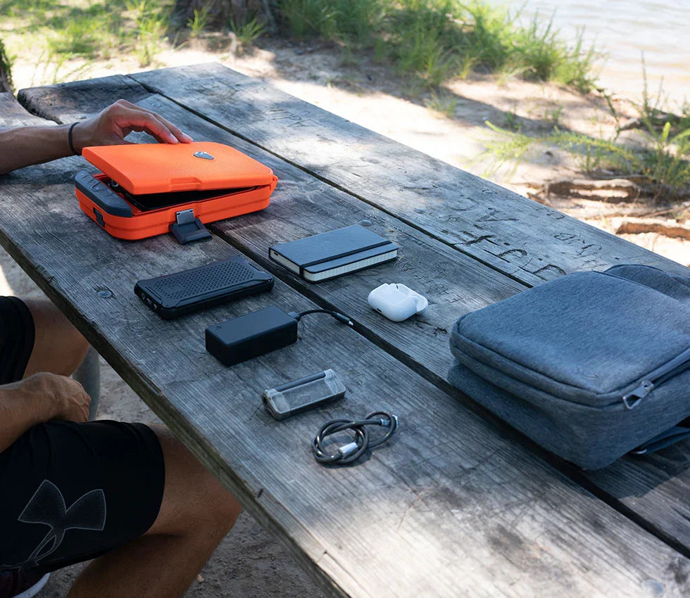 Outdoor picnic table with orange portable safe, tech accessories, and travel gear